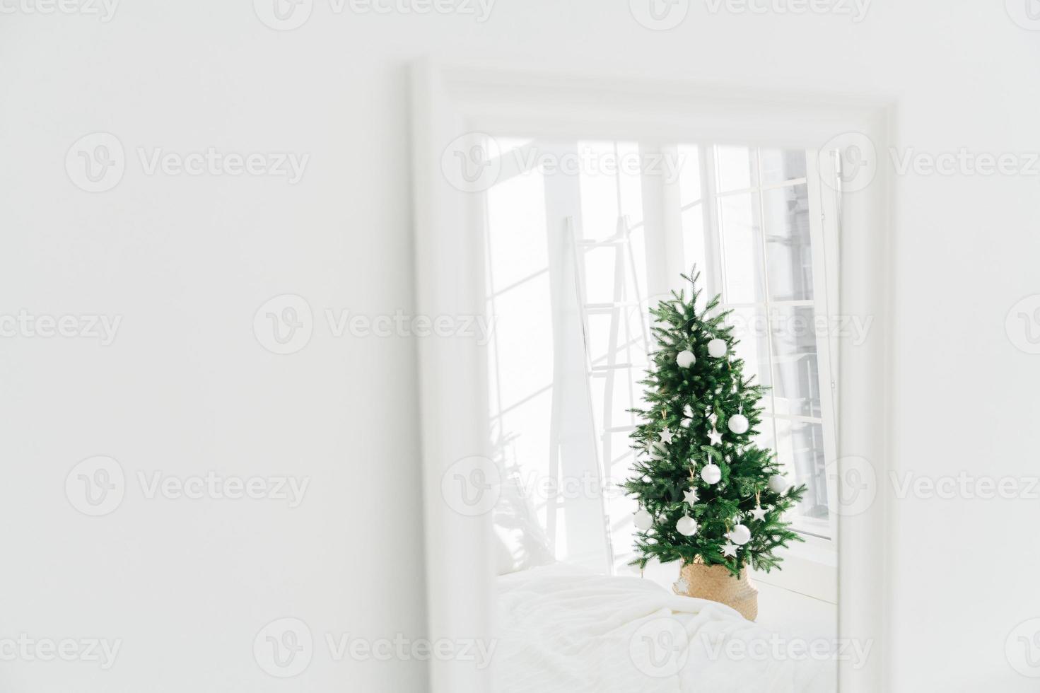 Decorated Christmas tree reflecting in mirror, white spacious room. Bedroom with New Year decoration. Modern classic style. Christmas eve at home photo