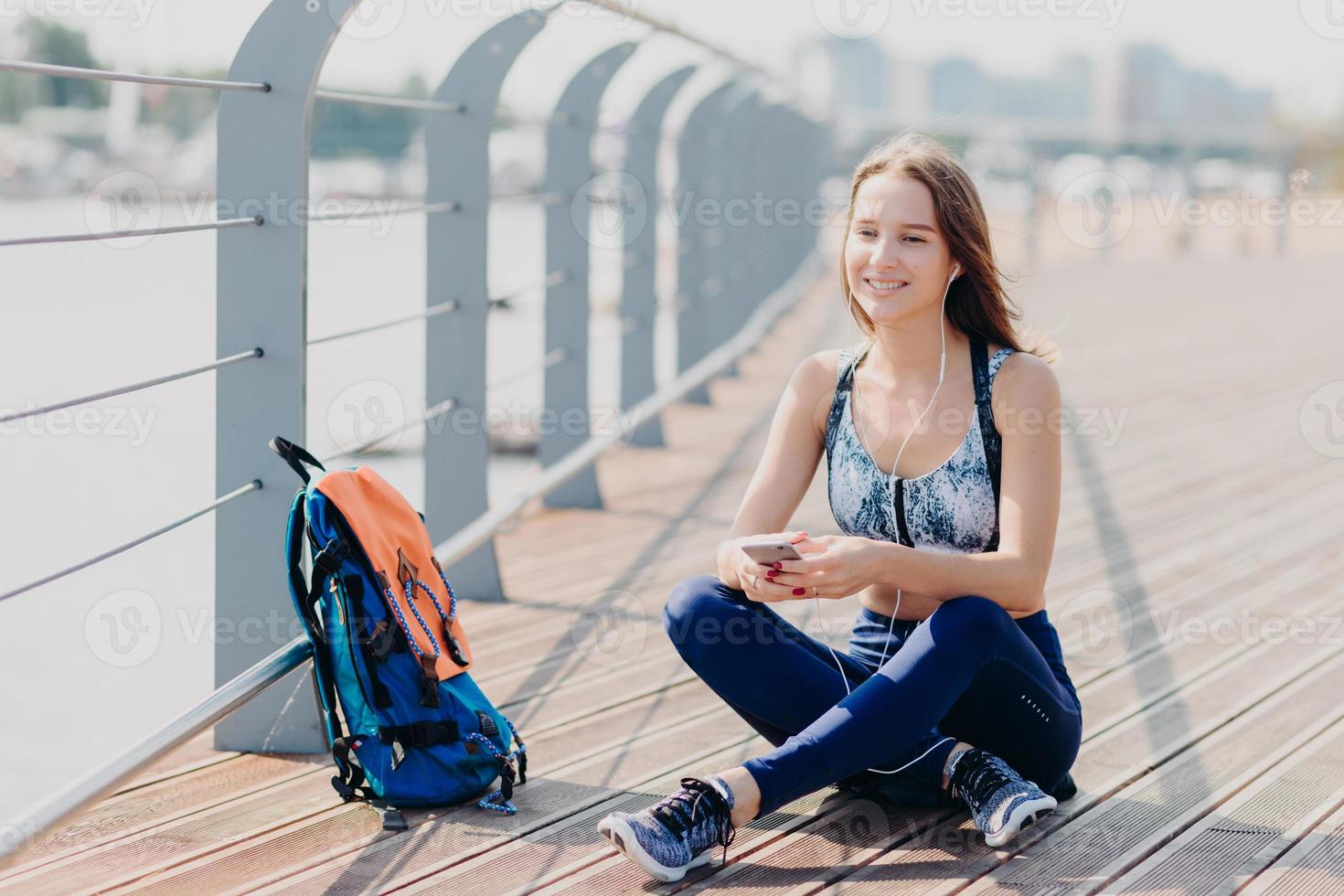 Photo of relaxed cheerful fitness girl in sporswear, sits crossd legs outdoor, rests after long walk, listens music with cellular and headphones, rucksack near. People, technology and recreation