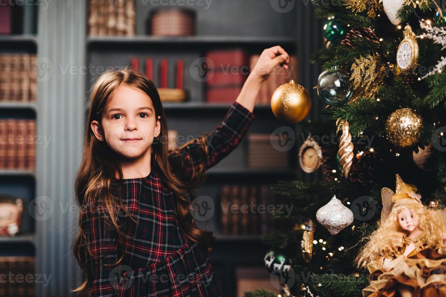 Small little kid decorates Christmas tree indoors, looks directly into camera, holds glass ball, being glad to have winter holidays. Little adorable girl wears dress decorates New Year tree alone photo