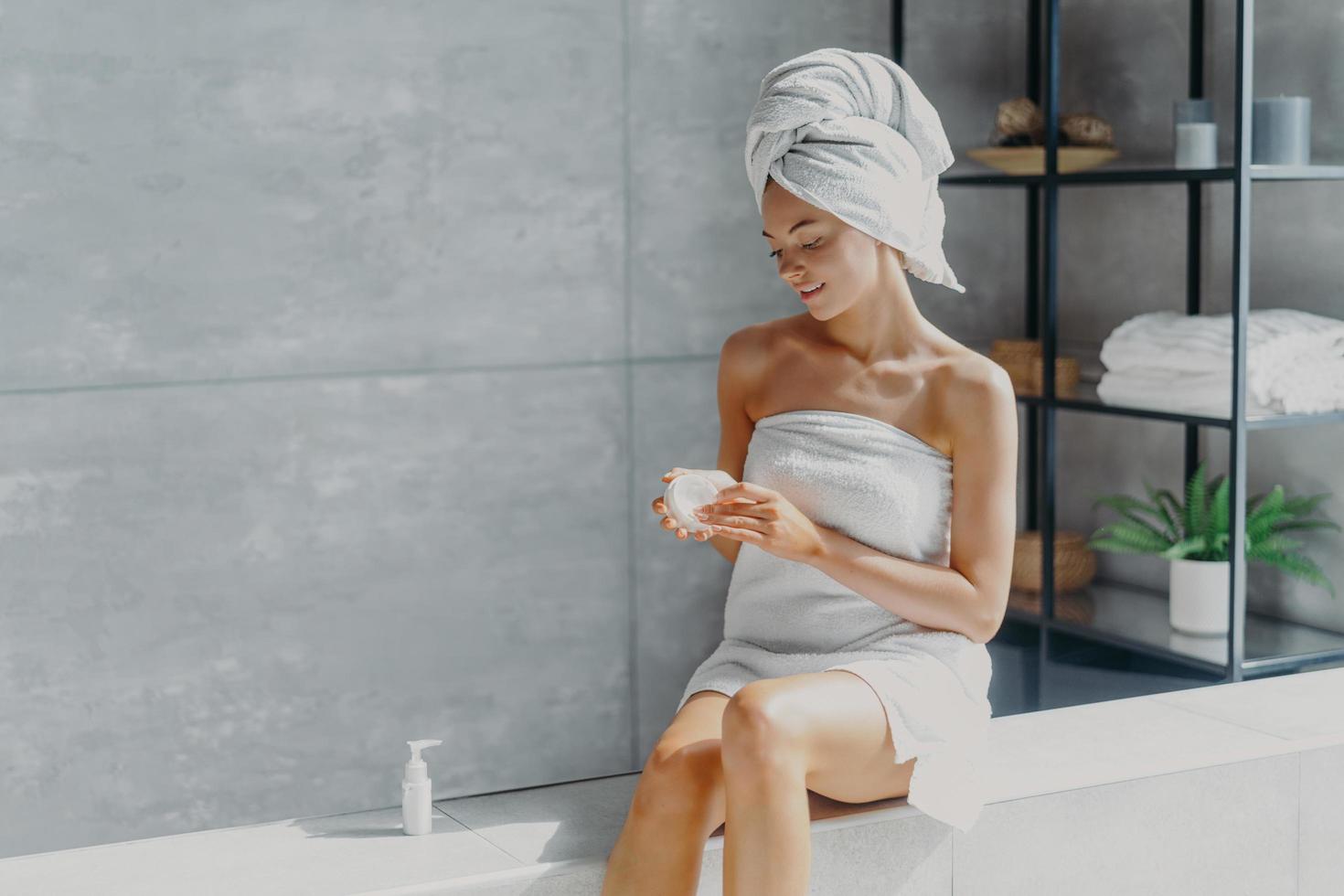 Pleased healthy woman wrapped in bath towel applies body cream after