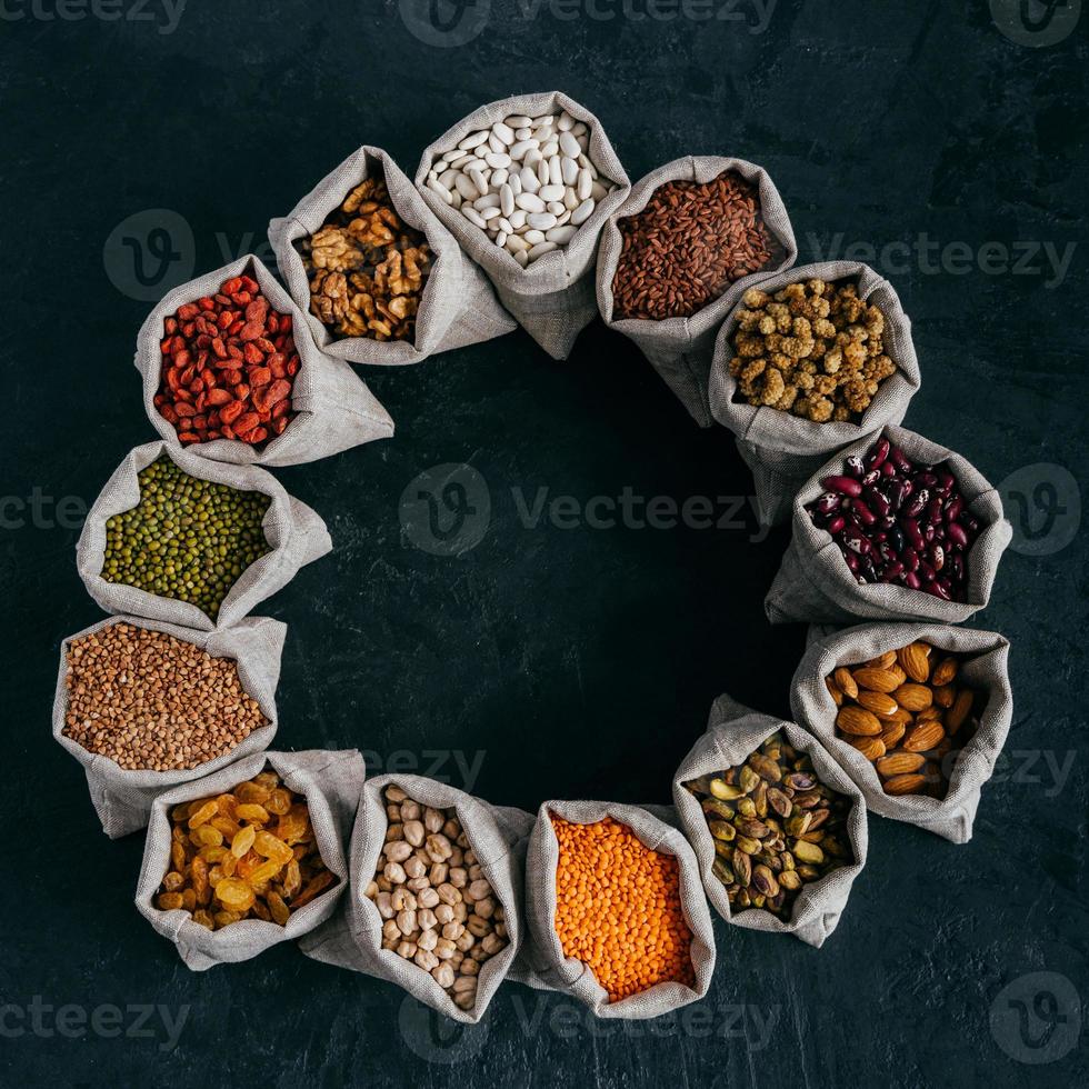 Assorted legumes and beans in sacks standing in circle, isolated over dark background with copy space in middle. Healthy eating concept photo