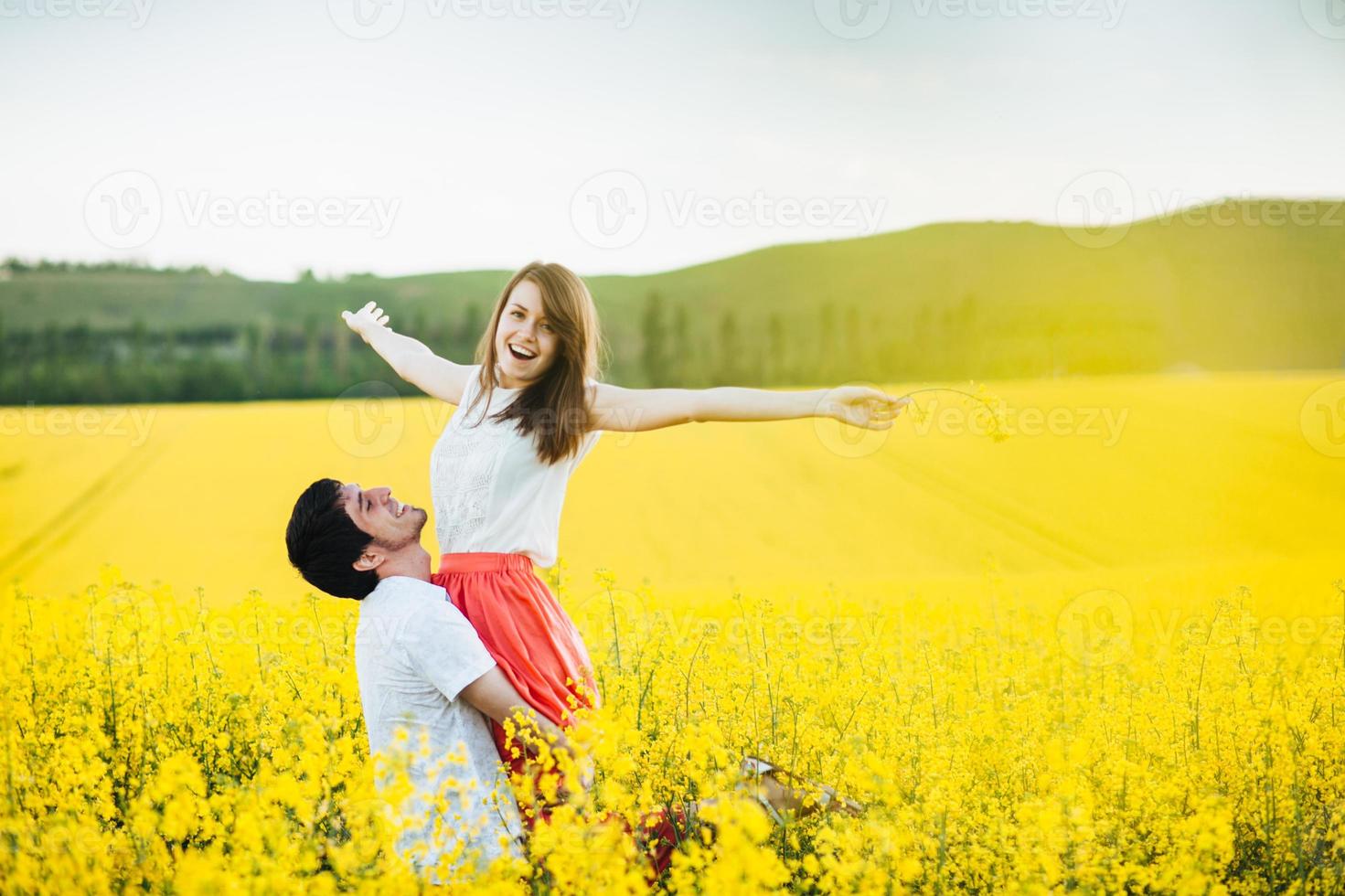 I feel free Joyful young female raises hands being on mans hands, pose together on yellow flower field during sunny summer weather. Romantic couple have fun outdoor. Relationships concept. photo