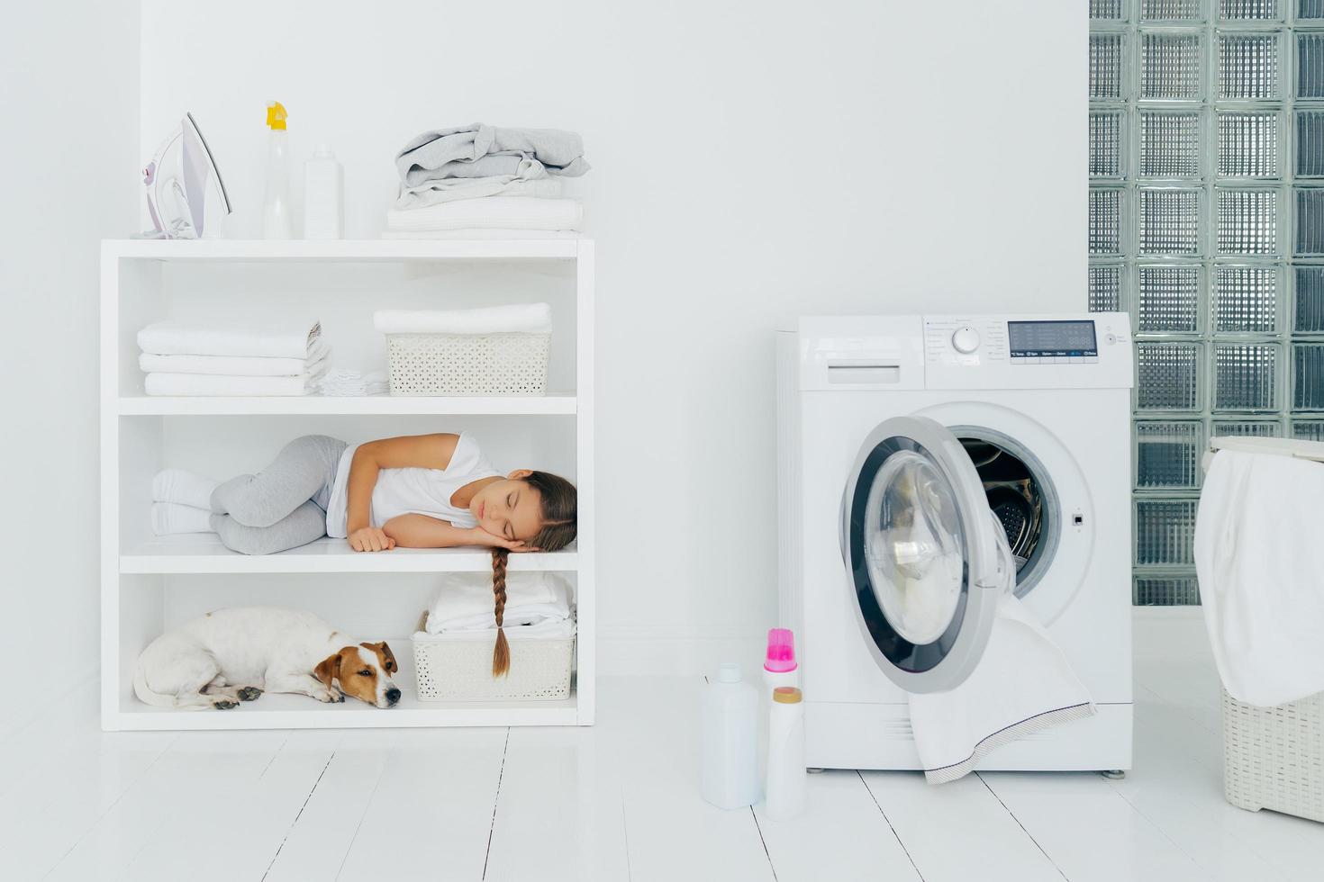 Tired female child rests on console shelf together with pet, does washing at home, sleeps in laundry room, opened washing machine with dirty towel inside. Childhood, cleanliness, family chores concept photo