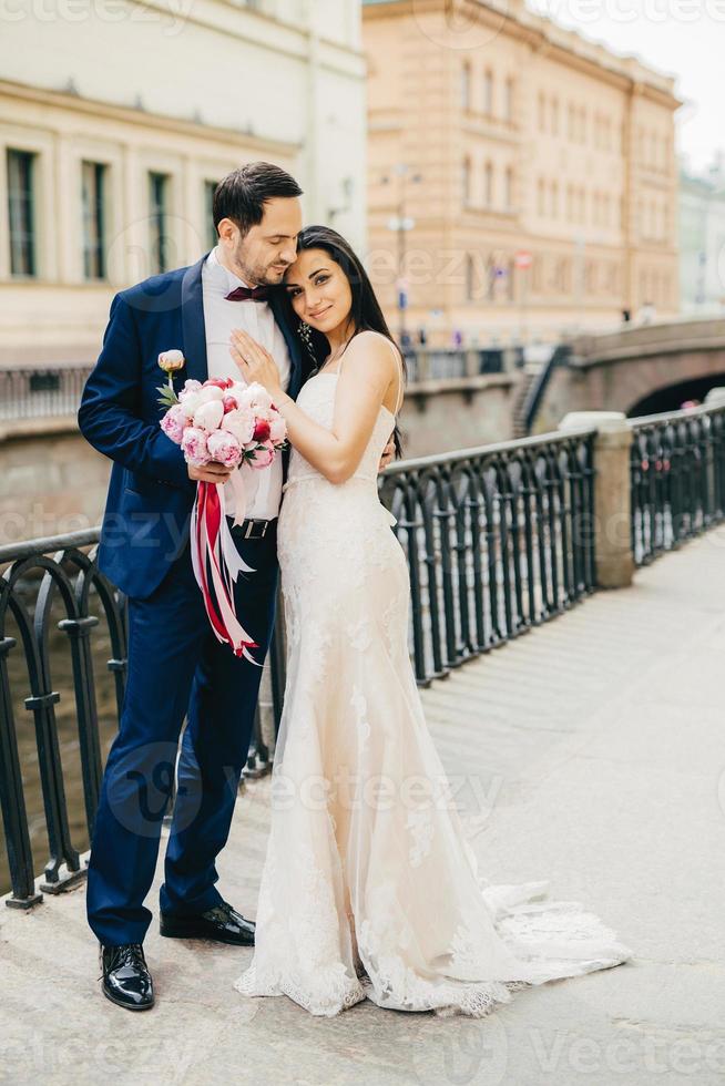 People, happiness and celebration concept. Beautiful young couple in love embrace each other at bridge, have model relationships, celebrate their wedding. Wife and husband have special occasion photo