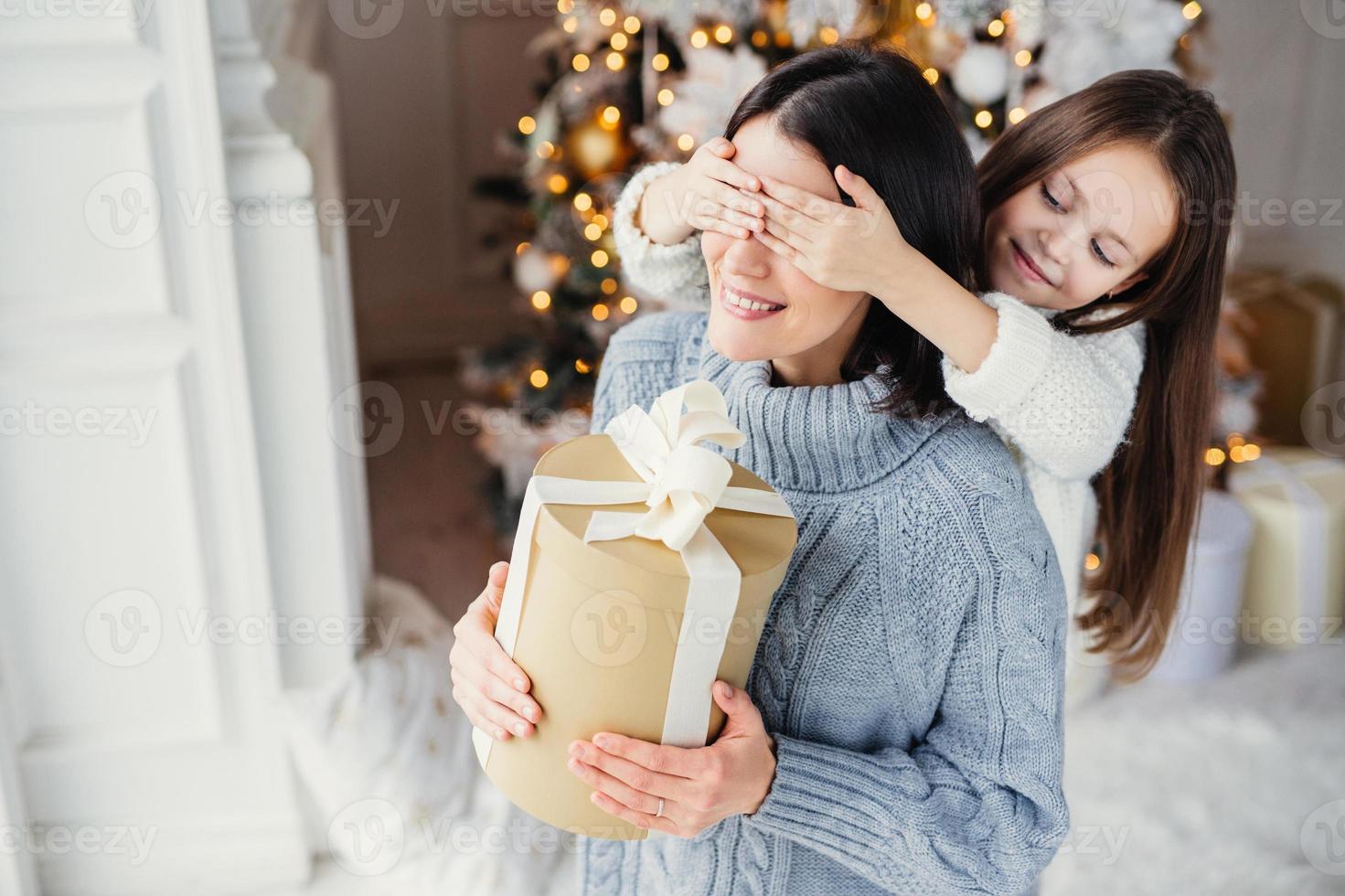 Small adorable female kid prepares surprise for her mother, closes eyes and gives wrapped present as stand at New Year background. Pretty female recieves gift from daughter. Surprisment concept photo