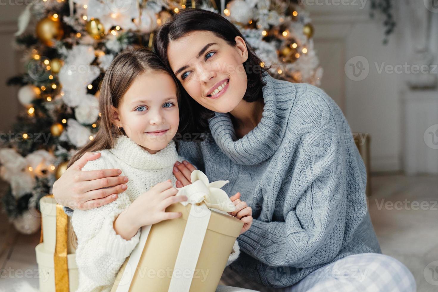 Cheerful brunette female leans at her daugter, embraces her, presents gift box, being in living room near decorated New Year tree. Glad family mother and daughter in warm sweaters celebrate Christmas photo