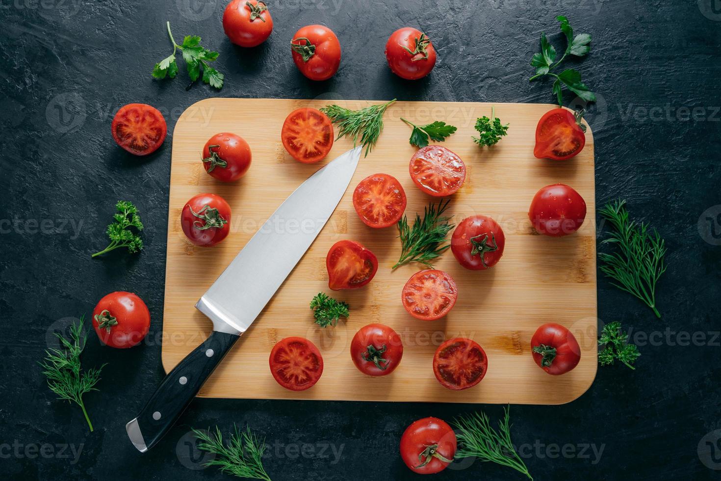 Top view of red sliced tomatoes on wooden chopping board. Sharp knife near. Green parsley and dill. Dark background. Preparing fresh vegetable salad photo