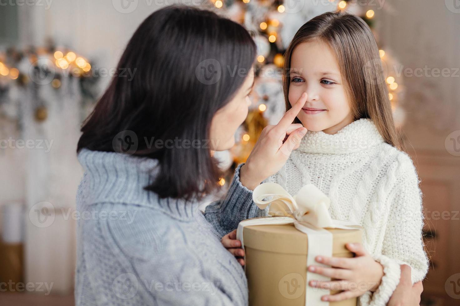 Affectionate mother gives present to her adorble little daughter, prepares surprise on Christmas, touch her nose, expresses great love. Family, celebration, presents, miracle, winter holidays concept photo