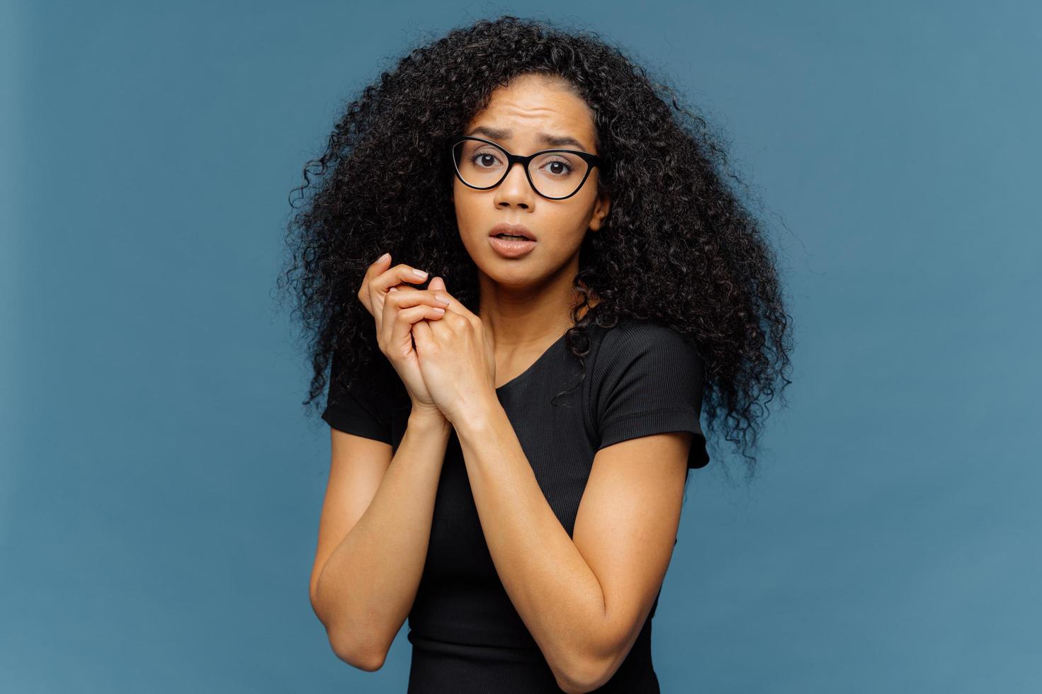 la foto de una mujer afroamericana avergonzada mantiene las manos juntas, mira nerviosamente a la cámara, tiene algunos problemas, usa una camiseta negra informal, se para contra el fondo azul. concepto de sentimientos negativos