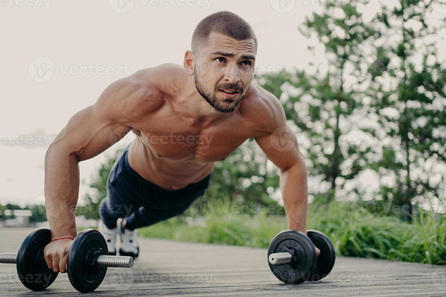 Young handsome bearded sportsman works out outdoor poses in plank with barbells, has strong body, muscular arms. Bodybuilder does exercises in open air, performs weight lifting, push up exercises photo