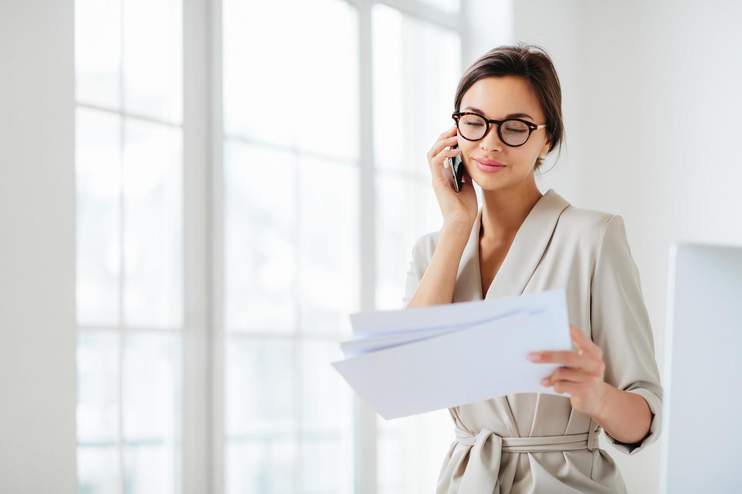 Horizontal shot of business lady discusses details of contract, works in office, concentrated on information from accountings, holds paper documents during telephone conversation, dressed formally photo