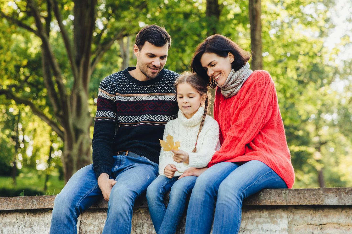 Adorable cute lovely small chld sits between her parents outdoors, holds leaf in hands, spend weekends together. Friendly young family enjoy togetherness, have happy expressions, sit on bridge photo