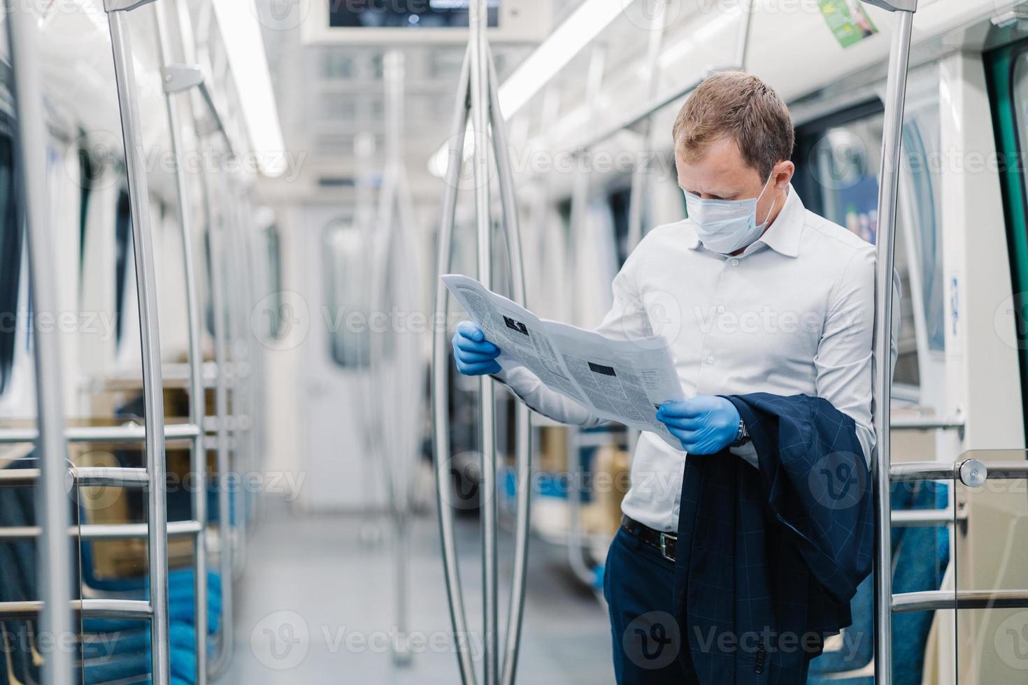 Horizontal shot of adult man wears formal clothes, protective medical mask and gloves, reads press, finds out news during virus outbreak, commutes to work in public transport. Coronavirus, Covid-19 photo