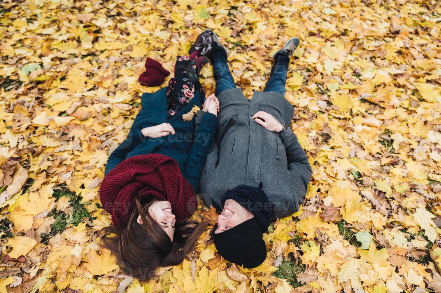 una pareja joven y romántica se acuesta en hojas amarillas en el parque, se toman de la mano, se miran con gran amor, disfrutan del día soleado de otoño. concepto de personas, relaciones, unión y romance. foto
