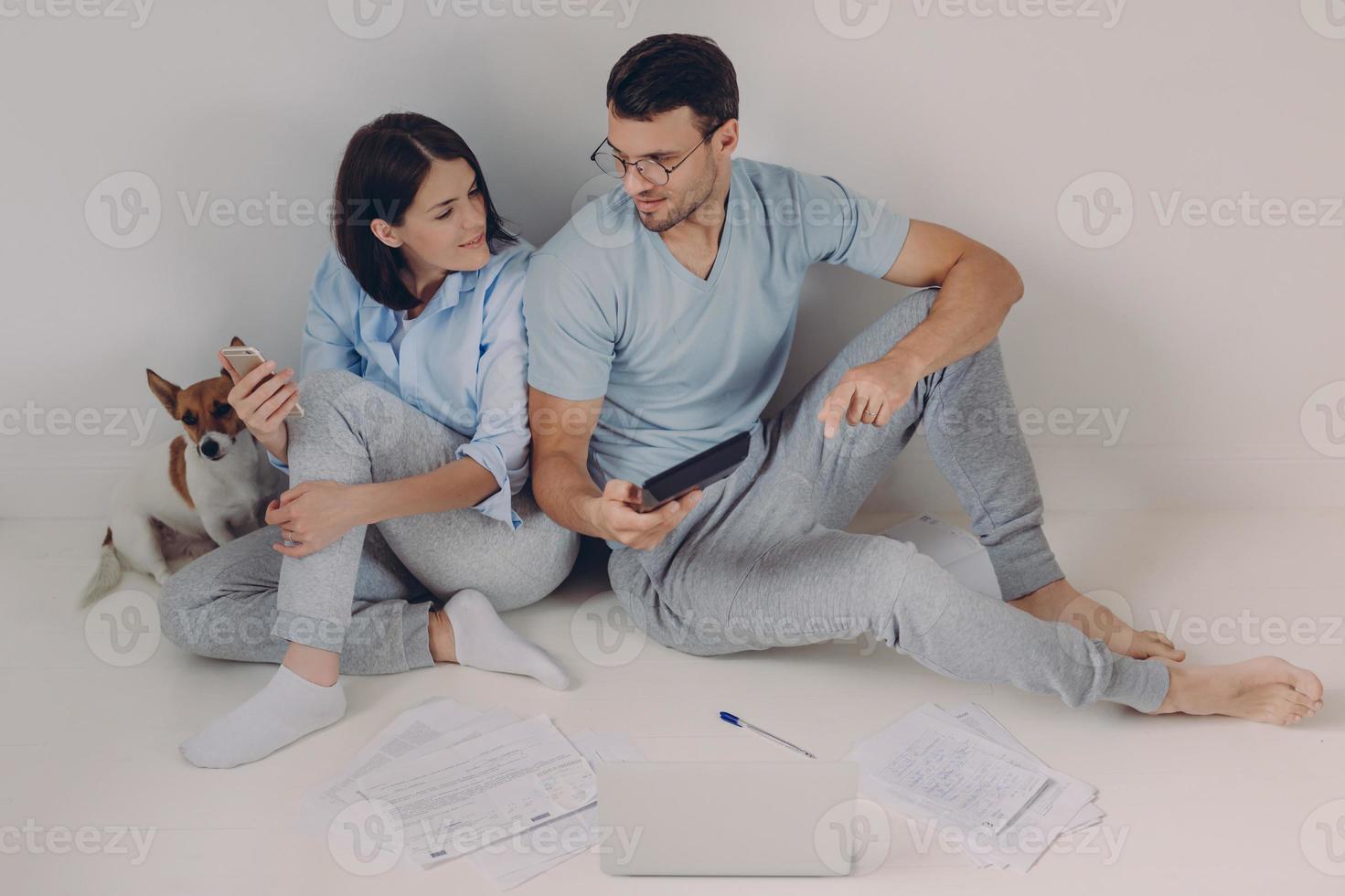 Cheerful young woman uses banking app on her mobile phone, his husband shows figures on calculator, surrounded with papers, have much work, solve economic problems, discuss something together photo