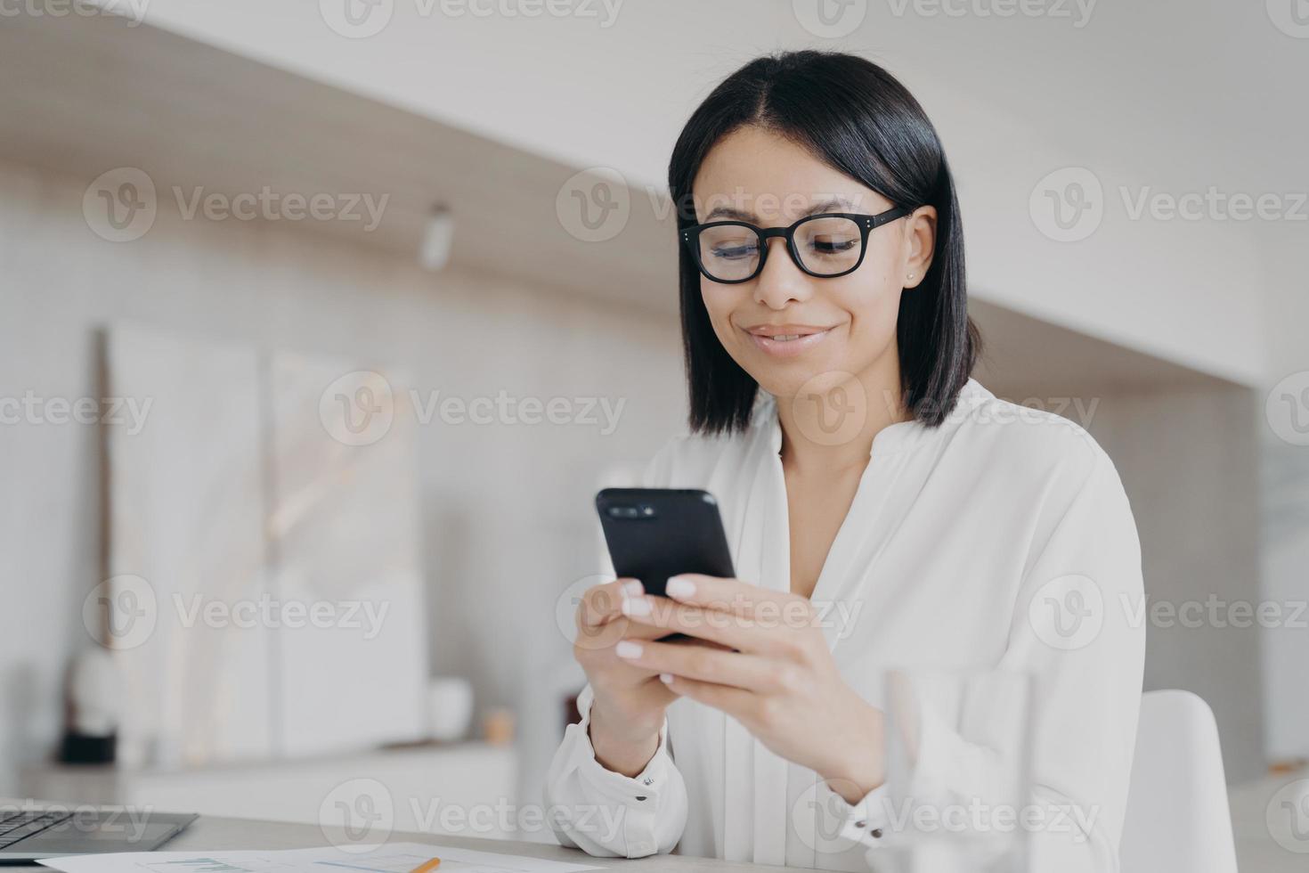 Businesswoman in glasses using phone mobile apps in office, reads good news message, chatting online photo