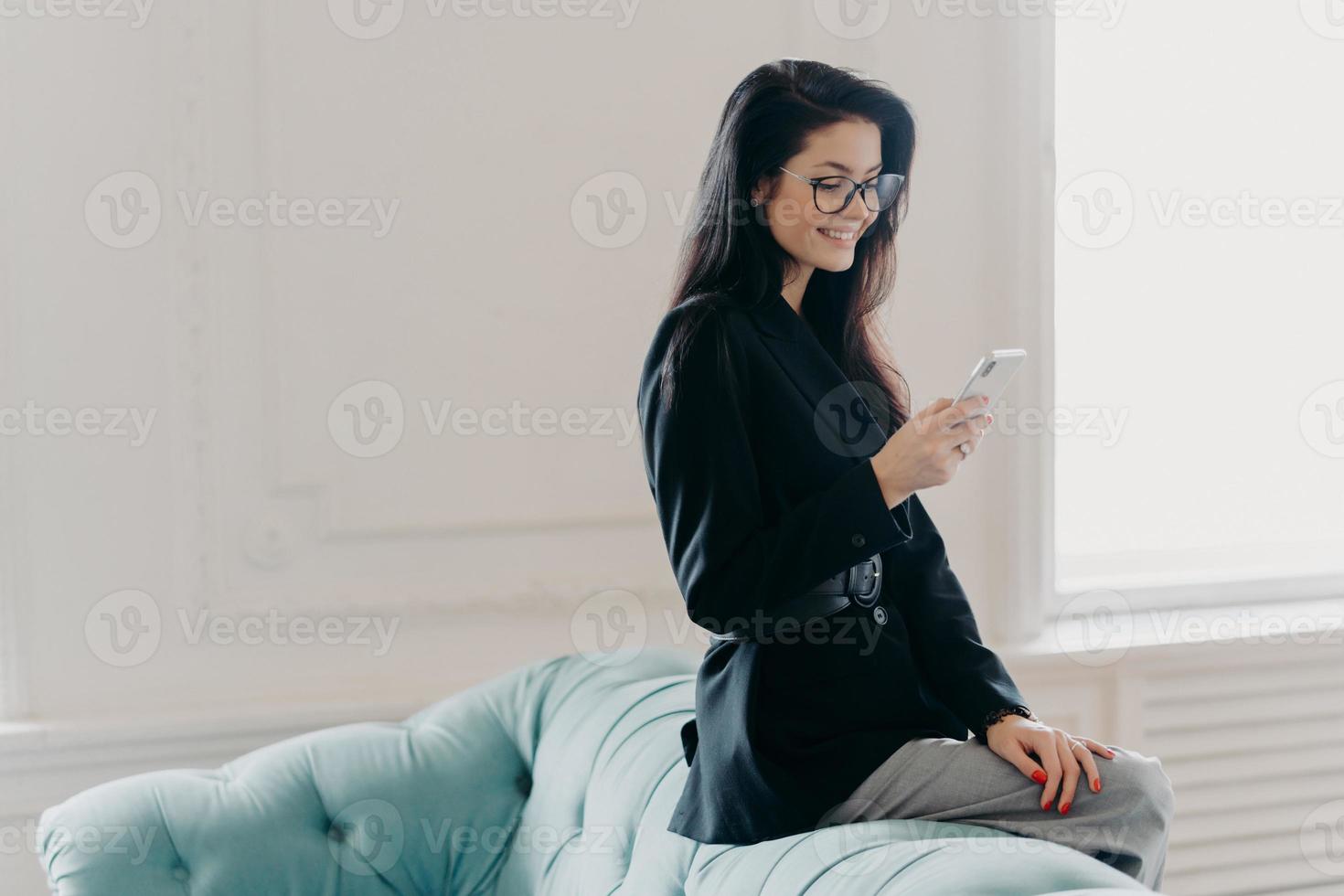 European entrepreneur wears corporate suit and spectacles, uses online banking application for transferring money, holds cellular, sends email, poses at back of comfortable sofa, uses free wifi photo