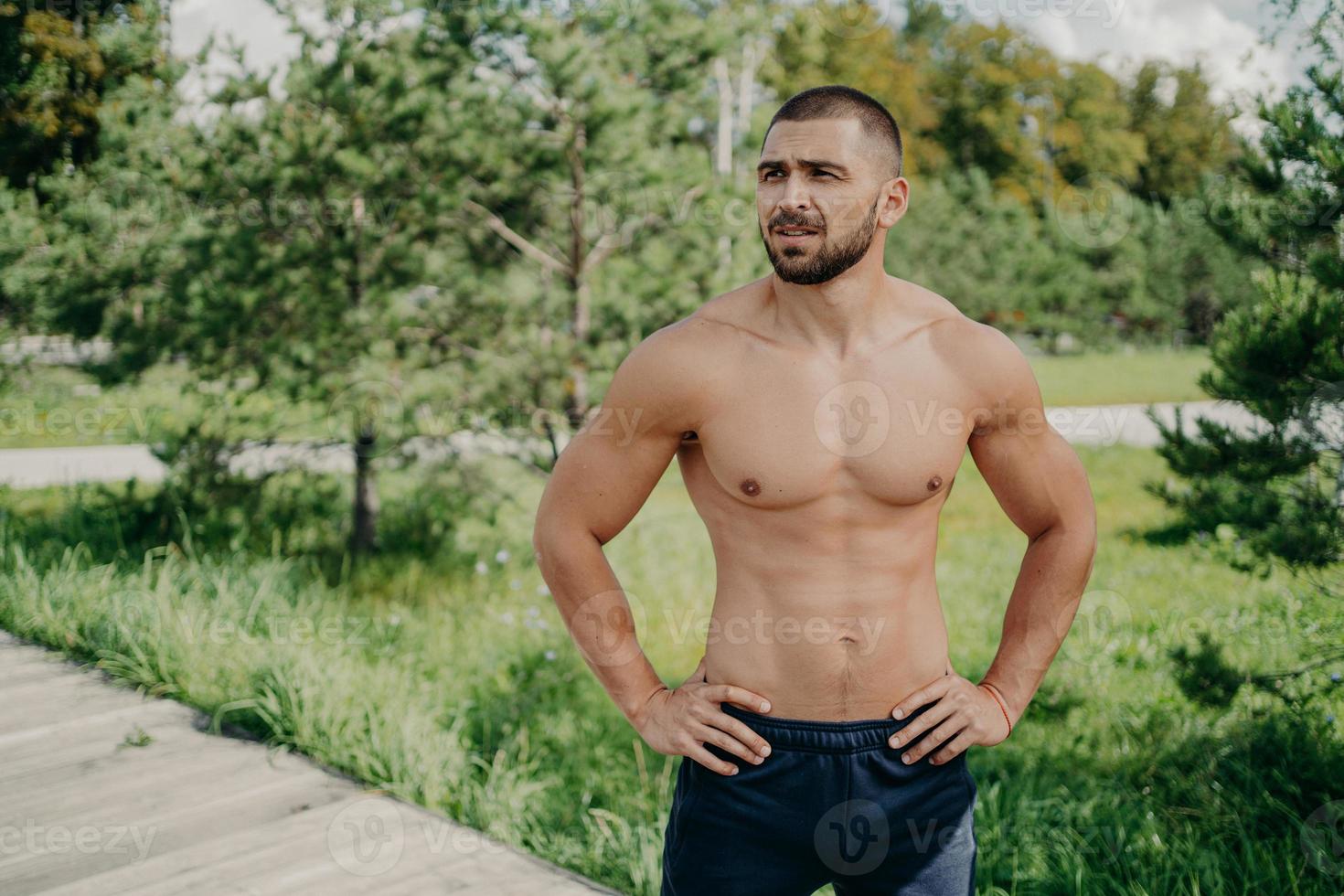 Horizontal shot of thoughtful shirtless sportsman keeps hands on waist, looks away demonstrates his muscular body, poses over nature background. Active bodybuilder goes in for sport, has daily workout photo