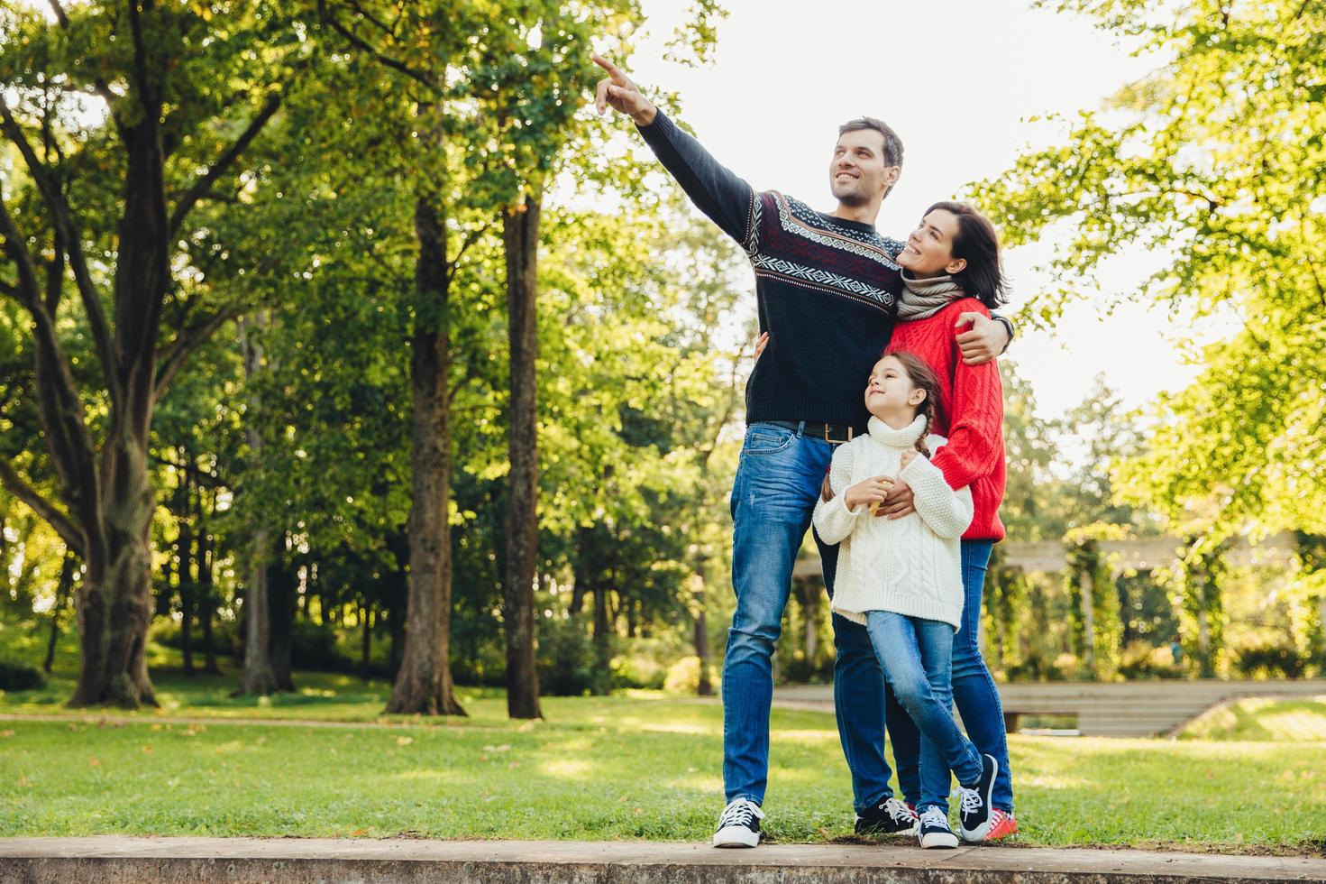 Image of happy joyful young family father, mother and little daughter play together in autumn park, countryside, enjoy nature outside. Attractive man shows something with hand daughter and wife photo