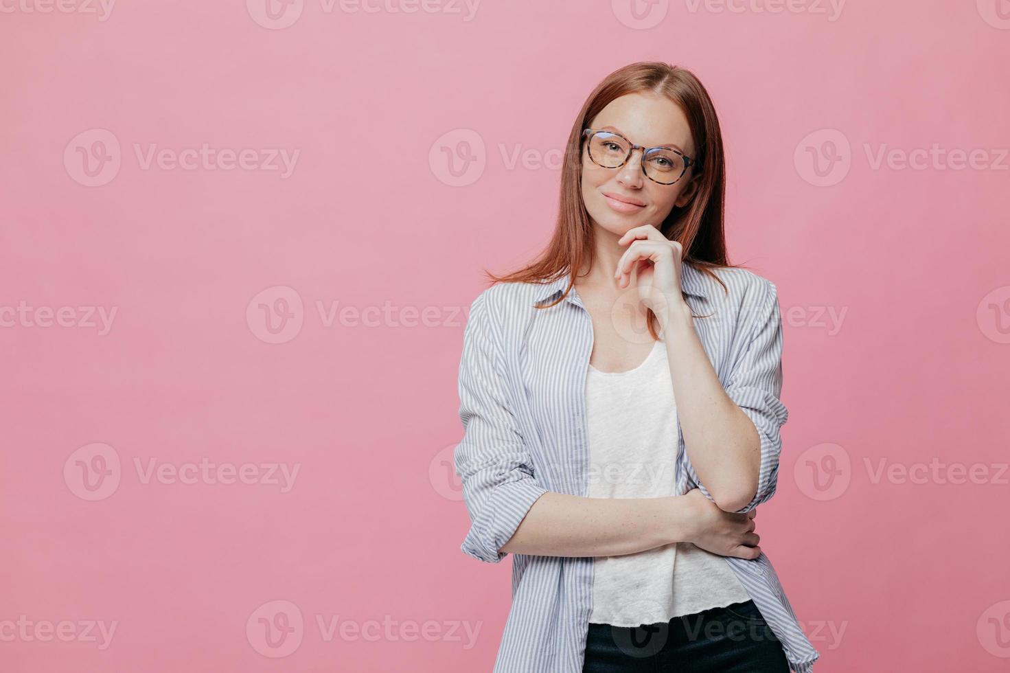 Indoor shot of thoughtful Caucasian woman keeps one hand under chin, dressed in elegant shirt, models over pink background, isolated over pink background with copy space for your promotion or slogan photo