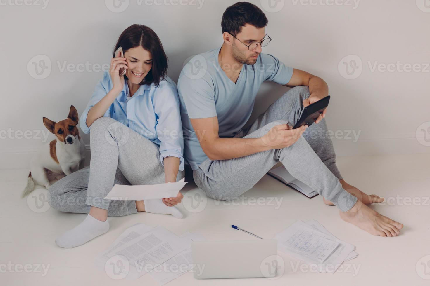 Young woman and man sit back to each other, do accounts together, make necessary calculations, surrounded with paper documents, sit on floor in empty room with dog. People and finances concept photo