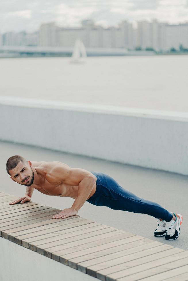 el hombre de ajuste flexible hace ejercicio de tablones en el paseo marítimo tiene el torso desnudo concentrado lejos se prepara para las prácticas de entrenamiento yoga vestido con ropa deportiva plantea trenes al aire libre por la mañana. estilo de vida deportivo foto