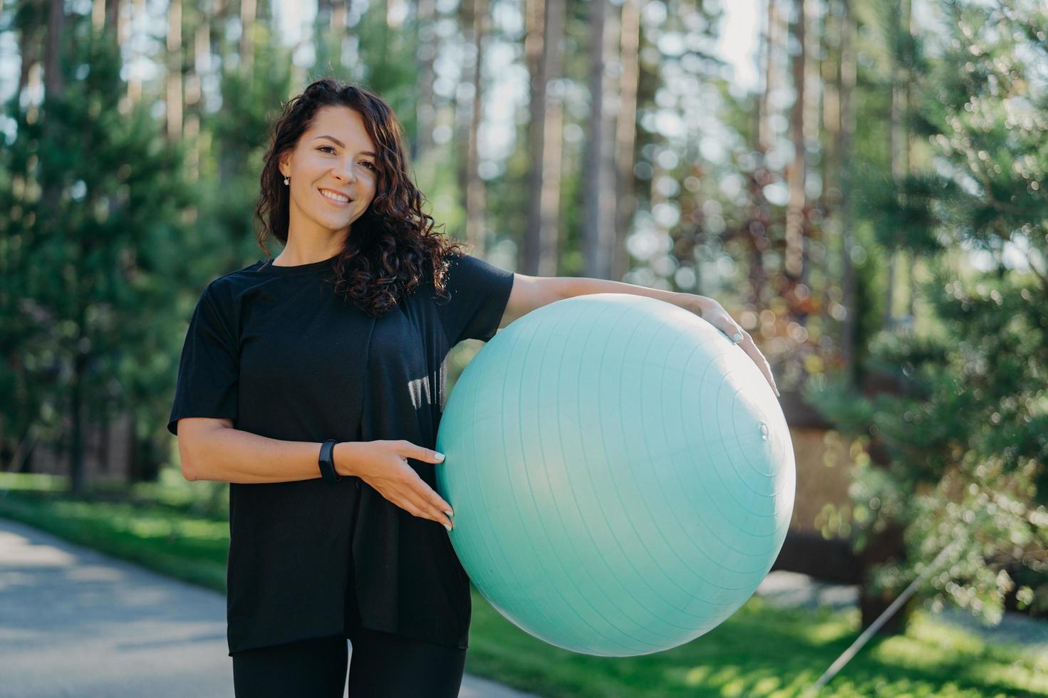 Positive brunette woman leads active lifestyle holds fitness ball poses against forest background dressed in black clothes has happy smile. Pretty female prepares for workout pilates does exercises photo