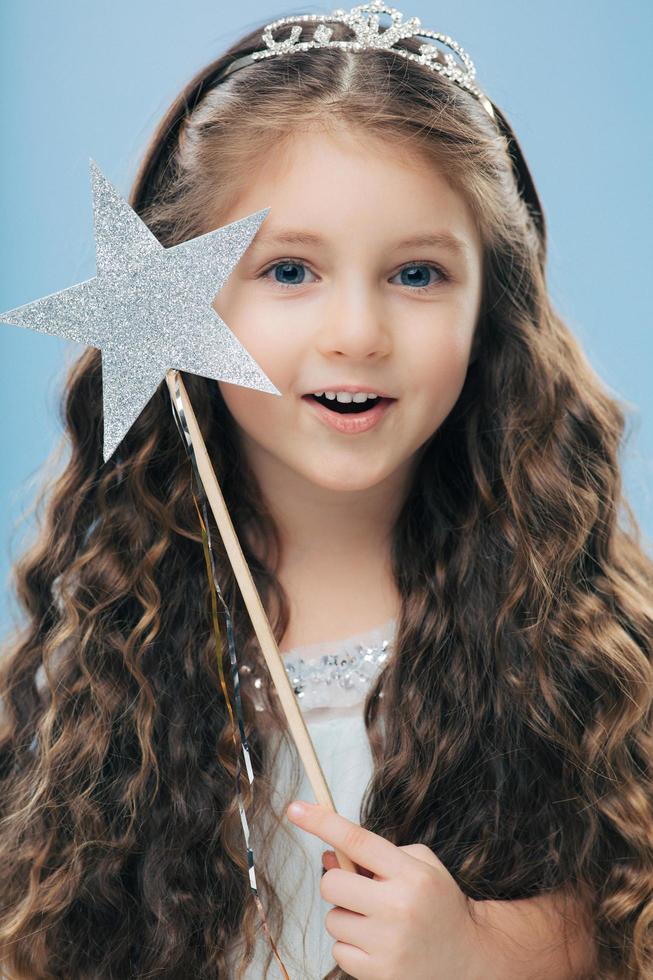 Close up shot of adorable blue eyed small European female princess has long wavy hair, wears crown, holds star magic wand, isolated over blue studio background. Vertical image. Childhood concept photo