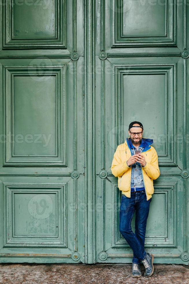 retrato completo de un hipster de moda con gorra de pie con las piernas cruzadas con un smartphone aislado sobre una antigua puerta verde. apuesto hombre barbudo leyendo sms en un teléfono celular genérico, enviando mensajes a amigos foto