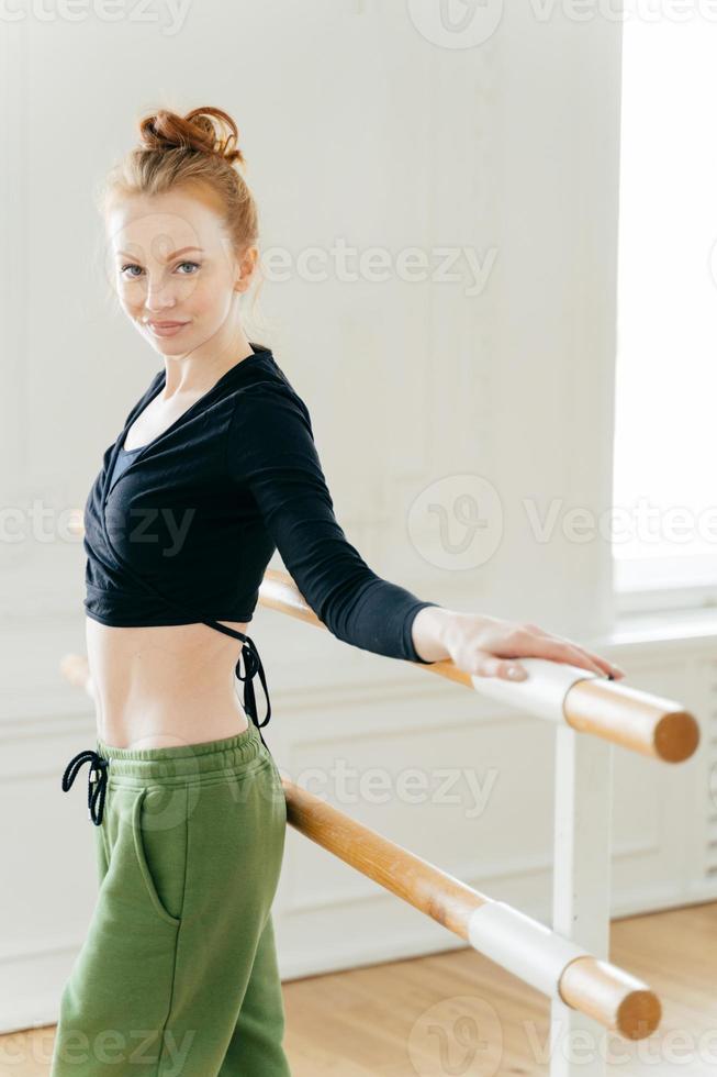 Half length shot of slim young female dancer stands back to barres, demonstrates dancing techniques, has determined look, shows fit figure, has make up, spends free time doing favourite hobby photo