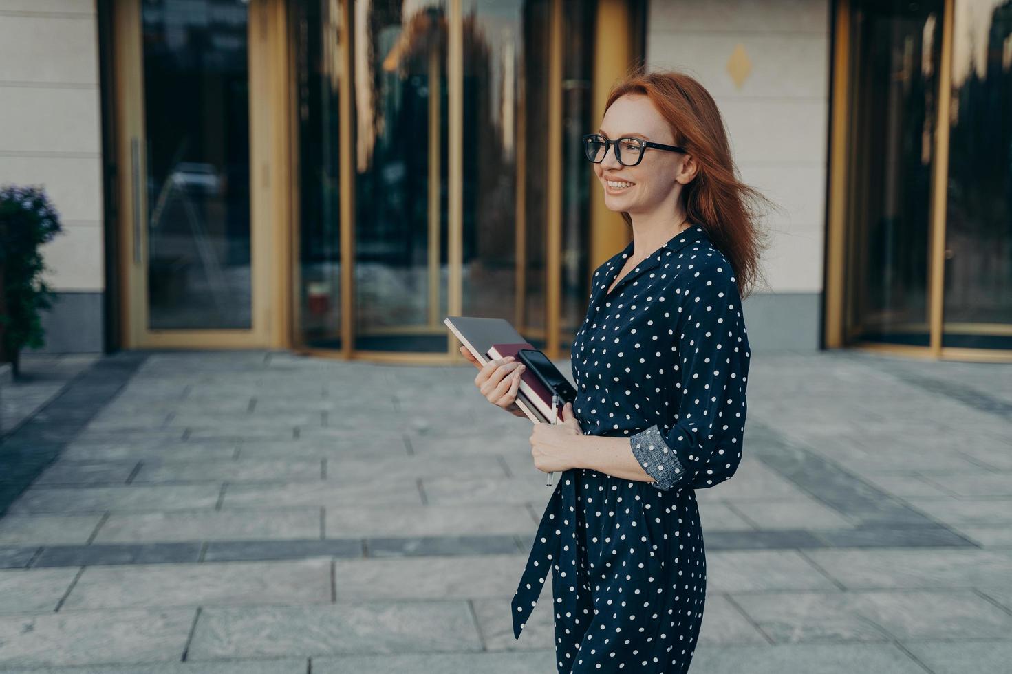 Skilled redhead businesswoman stands sideways has cheerful expression carries modern gadgets photo