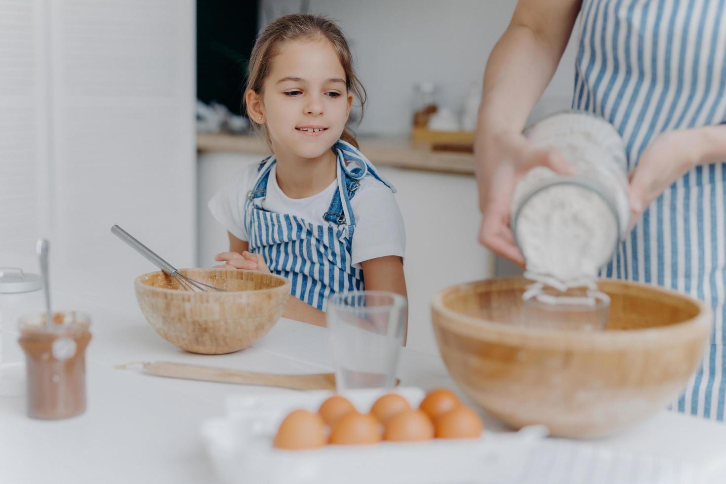 Curious little girl looks how mom prepares dough for pastry, learns to cook, gets culinary experience, wears apron. Faceless woman adds flour in bowl with other ingredients, pose in kitchen with child photo