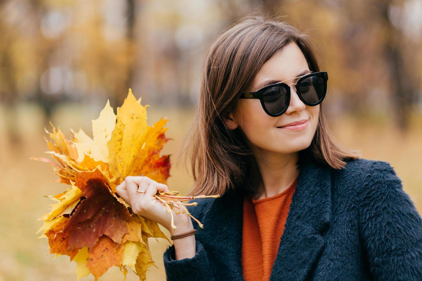 Close up shot of pretty woman with hair, wears sunglasses, has stroll during sunny day in autumn in park, carries yellow leaves, looks thoughtfully aside. People, season and lifestyle concept photo