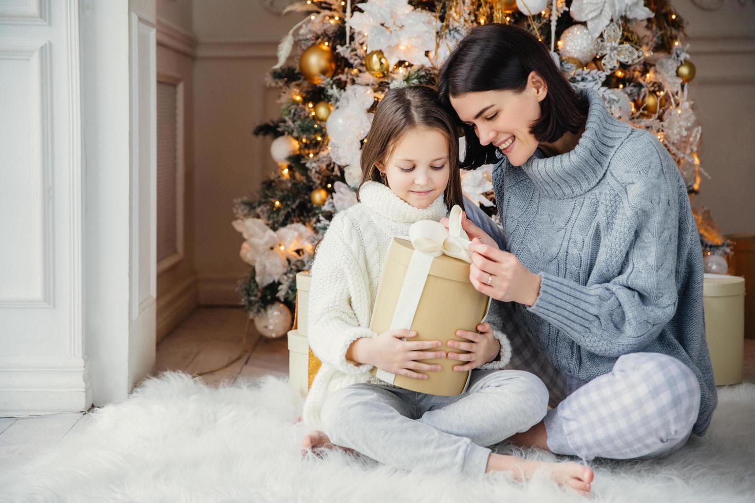 Indoor shot of adorable girl and her mother sit crossed legs, open wrapped present box, have intrigue what is there, sit against adorned New Year or Christmas tree, have sincere gentle smiles on faces photo
