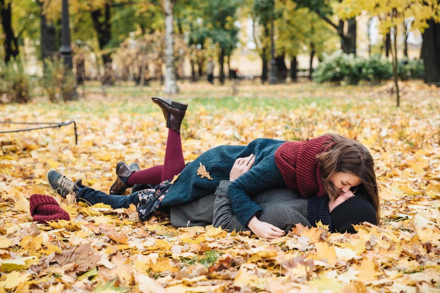 Boyfriend and girlfriend have romantic kiss as lie on yellow leaves in park, spend sunny autumn weekends together, have good mood. Couple in love enjoy togetherness outdoors. Season and people photo