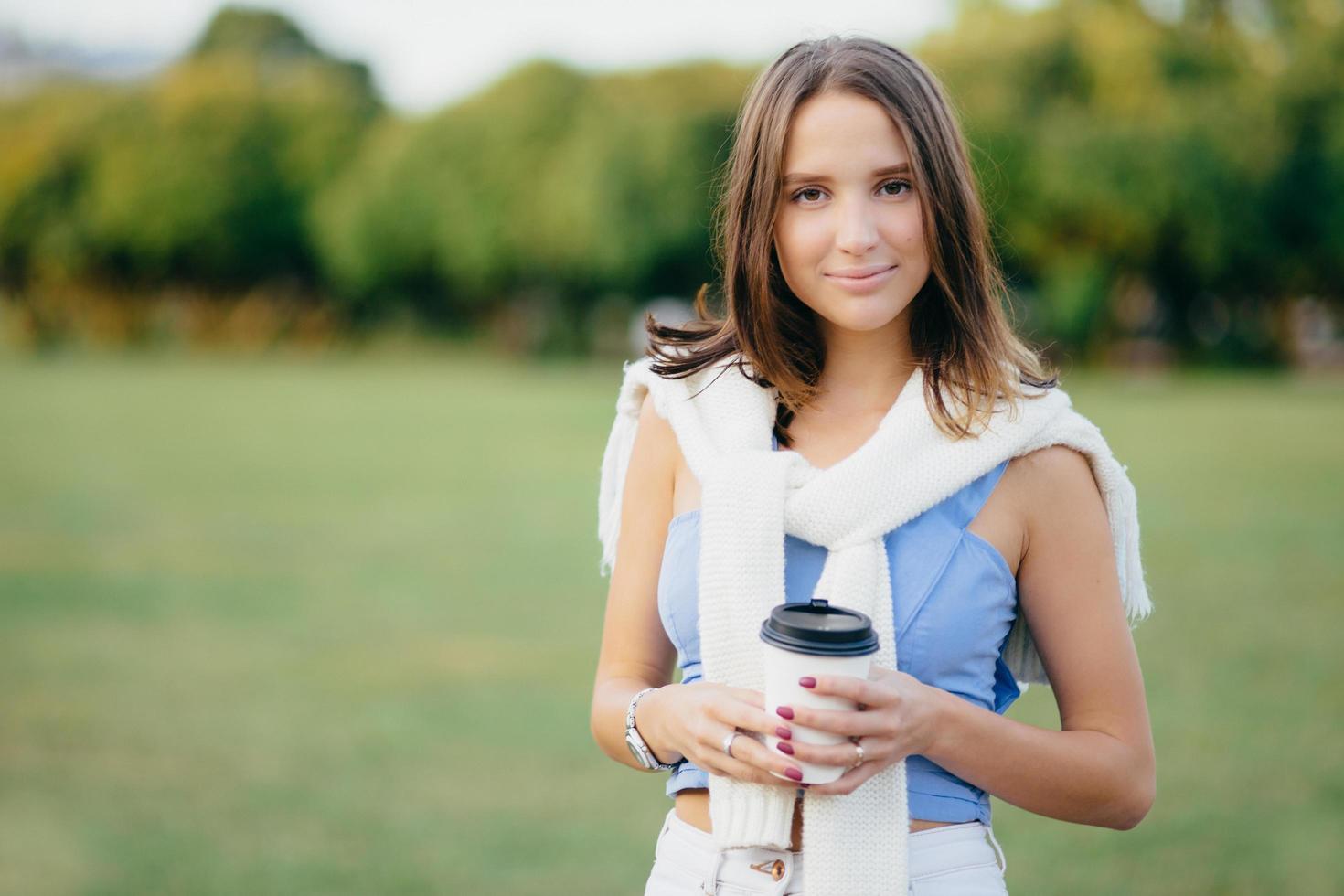 Photo of good looking woman with dark bobbed hairstyle, holds paper cup of coffee, has free time, spends weekend on nature, poses agaist green nature background with copy space for your text
