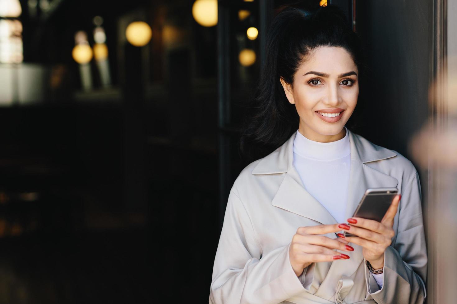 atractiva mujer de negocios morena con cabello oscuro atado en cola de caballo y maquillaje maravilloso sonriendo mostrando sus dientes blancos y perfectos esperando a su socio comercial usando un teléfono inteligente moderno foto