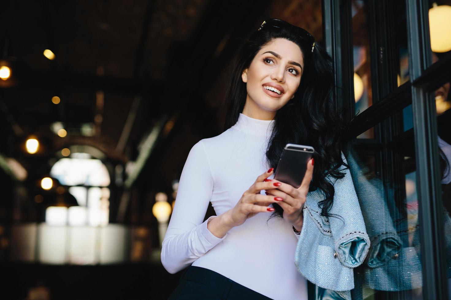 Dreamy stylish brunette lady with appealing appearance dressed elegantly holding mobile phone in hands watching photos remembering pleasant moments in her life. Woman with pensive expression