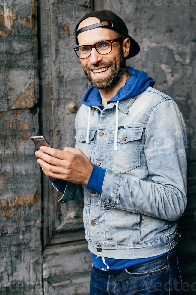 Stunning man with blue eyes, beard and broad smile wearing cap, denim shirt and eyeglasses holding smartphone isolated over old wall background. Pleased man in stylish clothes using cell phone photo