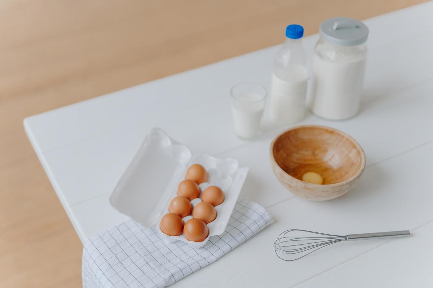 ingredientes para hornear en mesa blanca. huevos, leche y harina en recipientes. batidor para batir y hacer masa. proceso de horneado. hora de cocinar foto