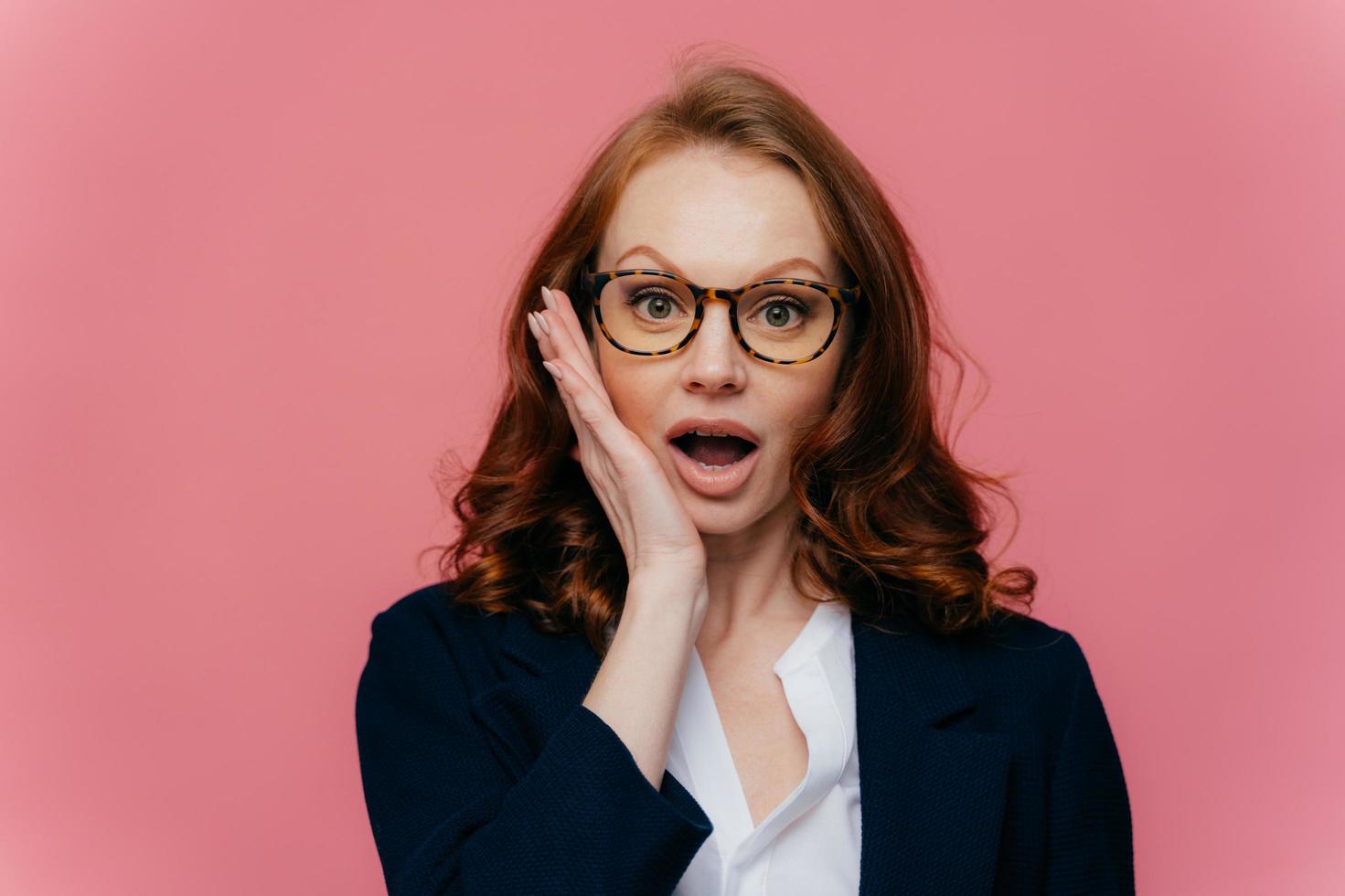 realmente de ninguna manera una mujer pelirroja sorprendida toca la mejilla, abre la boca ampliamente, mira sorprendentemente a la cámara, descubre noticias impactantes, usa anteojos, elegante traje formal, aislado en un fondo rosa foto