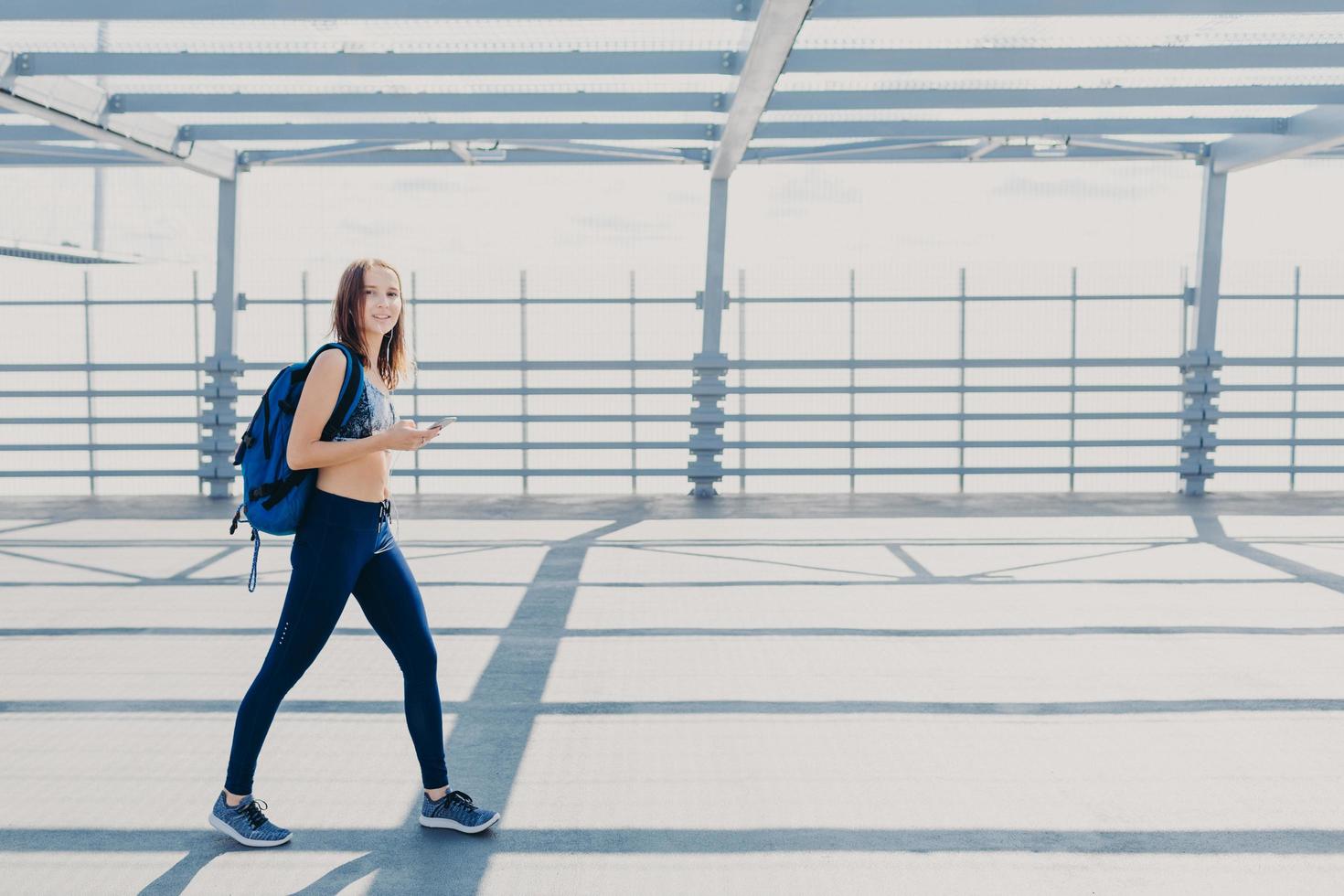 Outdoor shot of beautiful woman wth slim body, dressed in casual top and leggings, carries bag, going in gym, uses modern smart phone for listening music, copy space aside for your advertisement photo