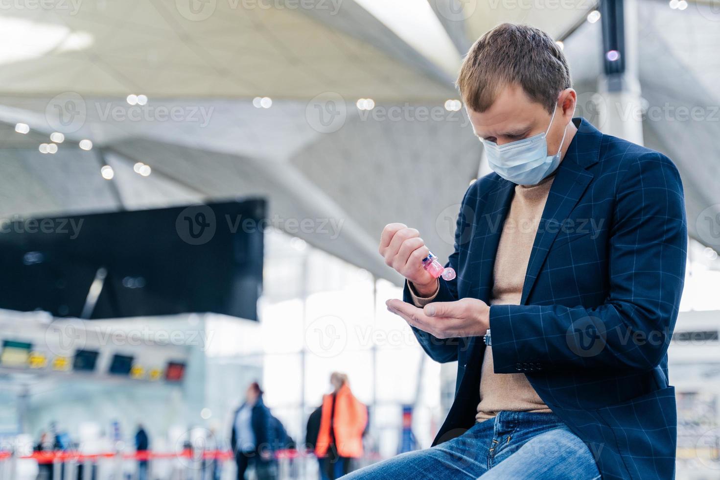 Photo of man in medical face mask, uses disinnfectant spray for hands, protects from coronavirus in crowded place, poses in airport, waits for flight. Health care concept. Sanitation, virus prevention