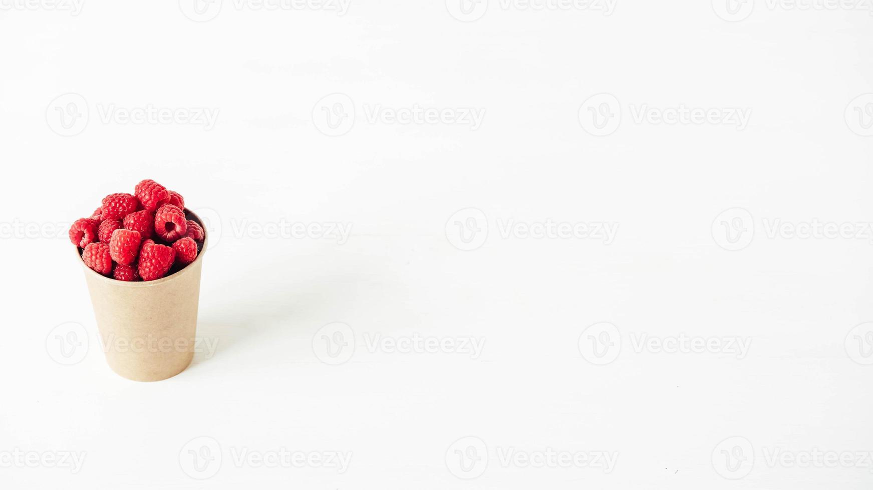 Fresh red raspberries in a paper cup on a white table background photo