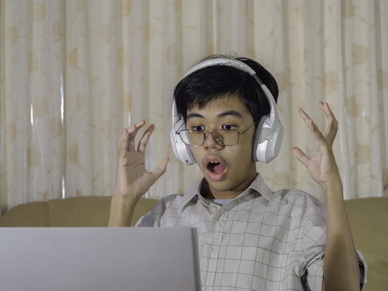 Teenage kid playing game console wearing headphone with raise hand after winning the game while sitting on sofa in living room. Online gaming concept. photo