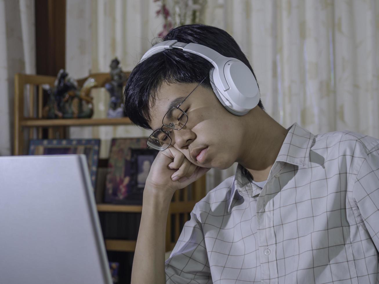 estudiante aburrido y soñoliento usando auriculares cansado de aprender a hacer la tarea sentado medio dormido en la pantalla de un portátil en su habitación. niño adolescente triste estudiando preparándose para la prueba de examen. foto