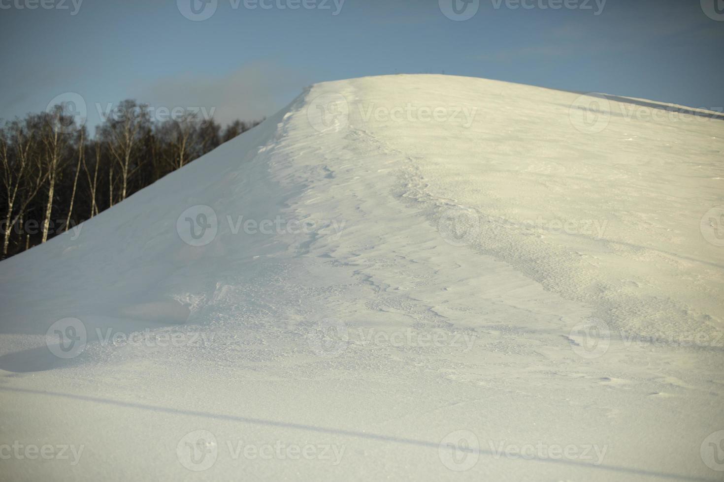 Mountain of snow. Snowy slope. Winter day. Sun is shining on hill. Natural object in park. photo