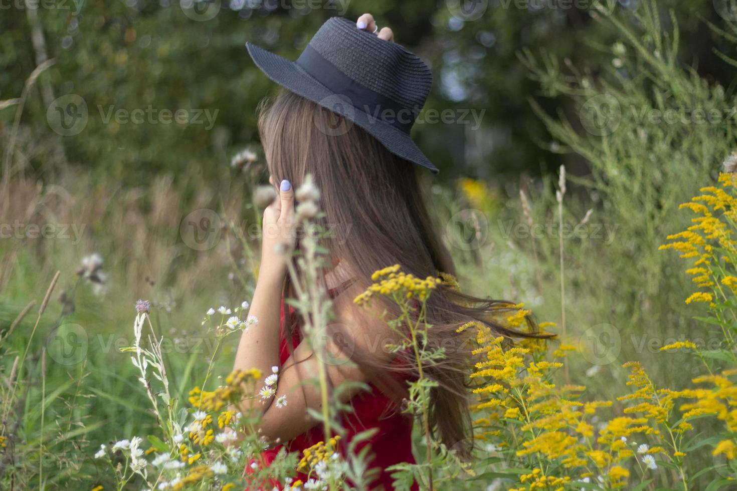 Girl in red dress stands in green clothes. Woman keeps black hat. Walk in park. photo