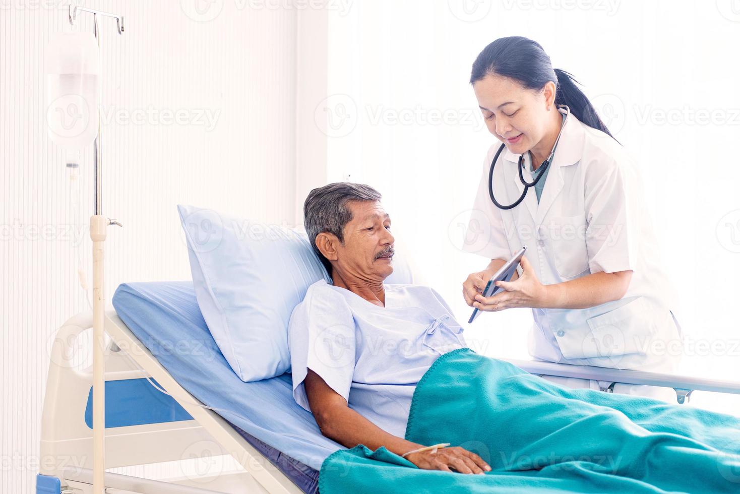 Asian woman professional doctor with clipboard visiting, talking, and diagnosing the old man patient lying in patient bed at hospital ward photo