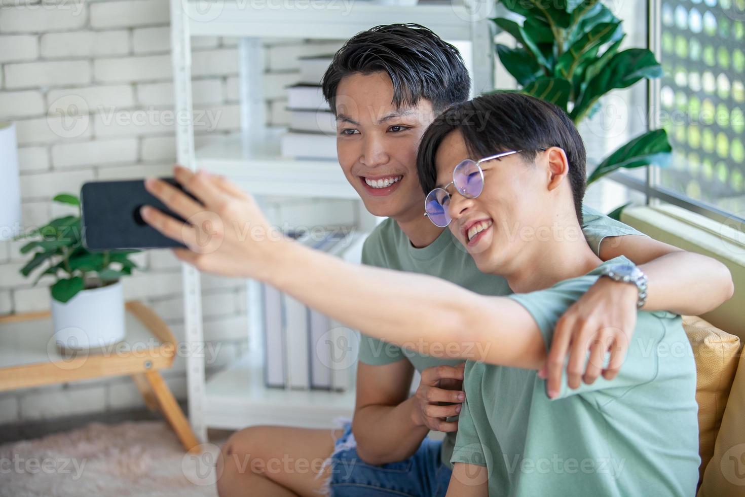 Happy Asian gay couple use mobile phone video call online or using an app on sofa, LGBTQ concept. photo